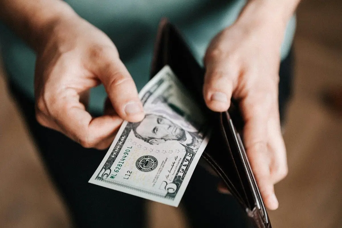 A person holds a five-dollar bill while retrieving it from a brown wallet, with a blurred background.