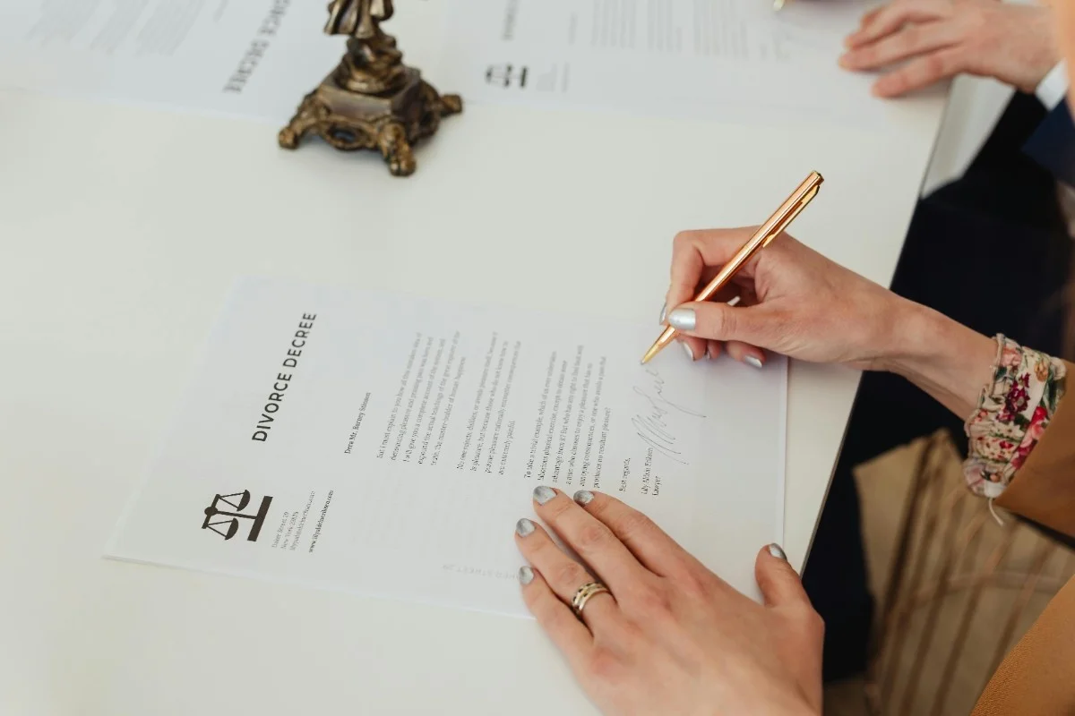 A hand with a gold pen signs a divorce decree document, with additional paperwork and a decorative scale figure in the background.