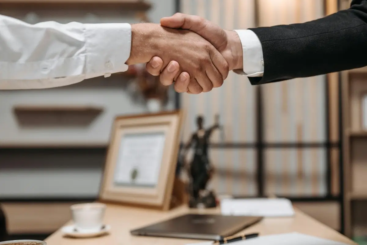 A close-up of two hands shaking, one in a white shirt and the other in a black suit, set against an office background.